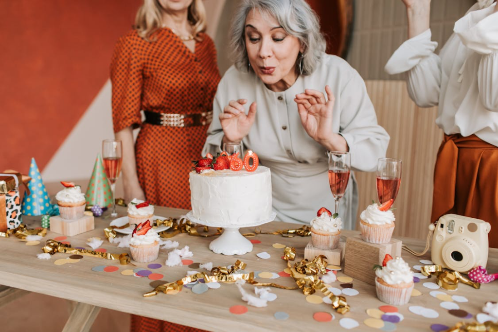 A joyful elderly woman with silver hair is blowing out candles shaped as '60' on a white frosted cake topped with strawberries. She is surrounded by friends dressed in stylish outfits, celebrating her milestone birthday. The table is adorned with cupcakes, champagne glasses, colorful confetti, and festive decorations, creating a warm and cheerful atmosphere.