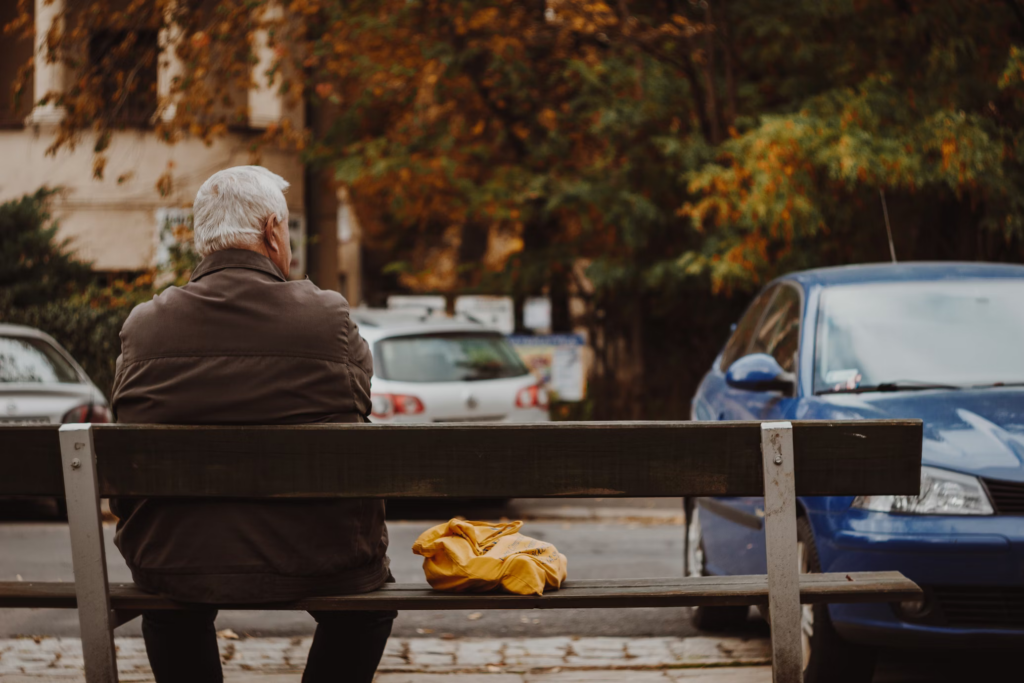 A senior man sits comfortably on a bench, with a car parked in front of him.