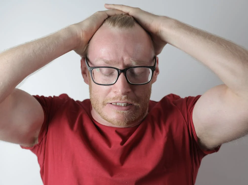 A man wearing glasses and a red shirt is holding his head, indicating concerns about mood changes and memory problems.