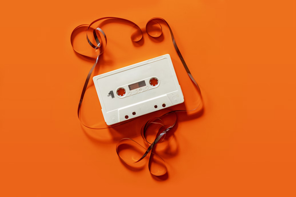 A vibrant orange background featuring a classic cassette tape with a colorful ribbon intertwined beside it.