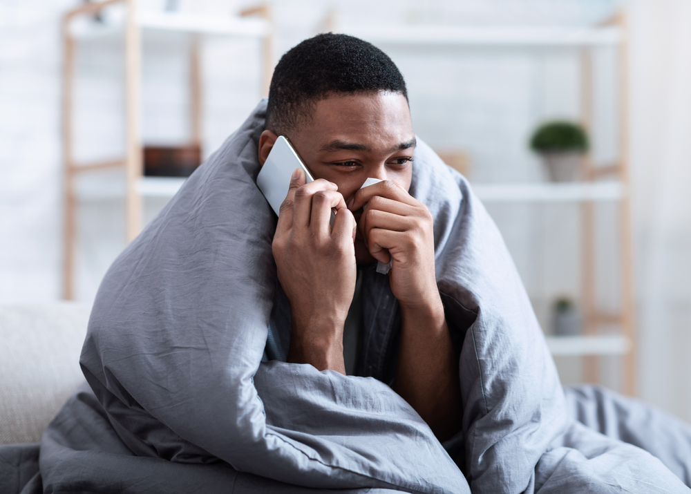 Ill Black Man Calling Doctor Having Runny Nose Suffering From Cold And Rhinitis Sitting On Sofa At Home. Sick African Guy Talking On Phone Feeling Unwell. Symptoms. Influenza Symptoms