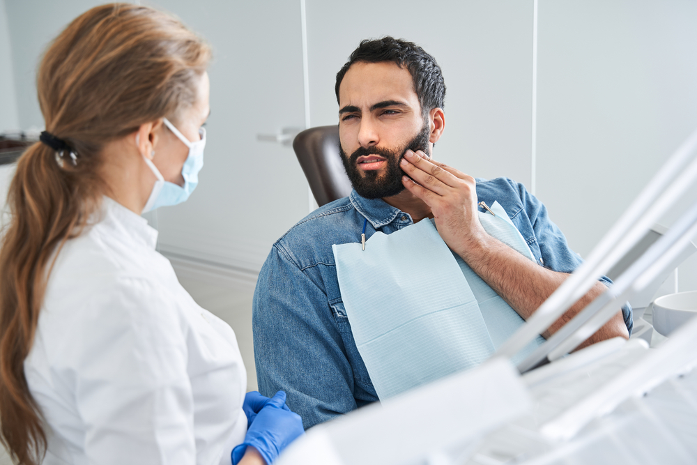 Feeling bad. Young multiracial man having toothache, sitting in a dental chair at the clinic. Bearded male patient with terrible toothache visiting dentist. Stock photo